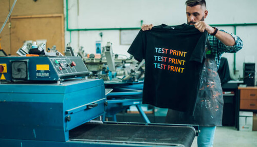 Low-investment business ideas A person holding a t-shirt with a test print design in a screen printing workshop, illustrating a low-investment business idea in the custom printing industry.