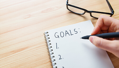 Set goals A close-up view of a hand writing a numbered list under the heading "goals:" in a spiral notebook, with reading glasses nearby. This symbolizes planning and setting objectives crucial for working in a business.