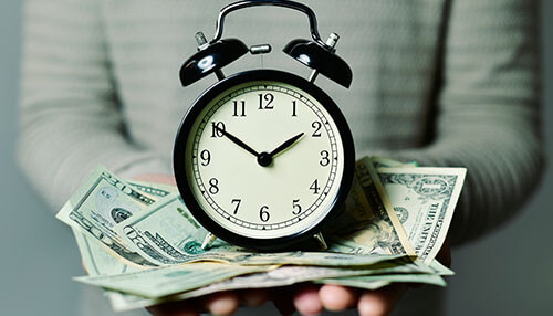 Both time and money are saved - tycoonstory | tycoonstory media An alarm clock and dollar bills held by a person, symbols of time and cost efficiency with recruitment process outsourcing companies.