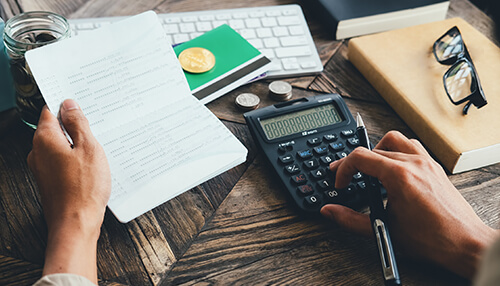 Checking the bank statements A person reviewing a long receipt while calculating expenses, dealing with late payments that impact their budget, shown with coins, documents, and a calculator on a wooden desk.