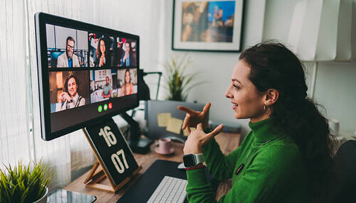 Easy to attend Woman participating in virtual events on a computer, smiling and gesturing while engaging with multiple people in an online video meeting.