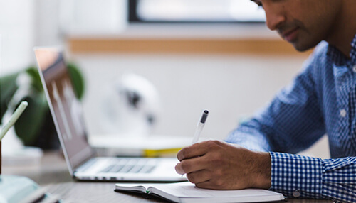 How to develop self-discipline Man writing in a notebook next to a laptop, exemplifying self-discipline and focus in a productive workspace