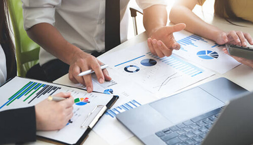 Managed services salesforce offerings for businesses A close-up of a business team’s hands as they analyze printed financial reports featuring bar charts, pie graphs, and line trends. A laptop and pens are on the table, reflecting a collaborative data review session.