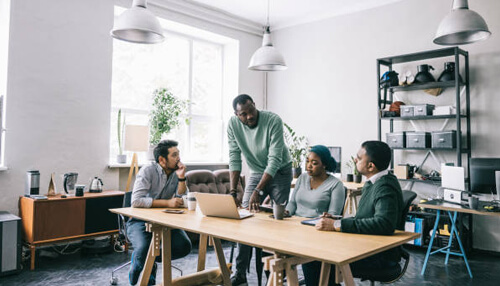 Project manager A diverse group of four professionals in a modern, light-filled office collaborating around a large wooden table with a laptop, reflecting a productive team environment.