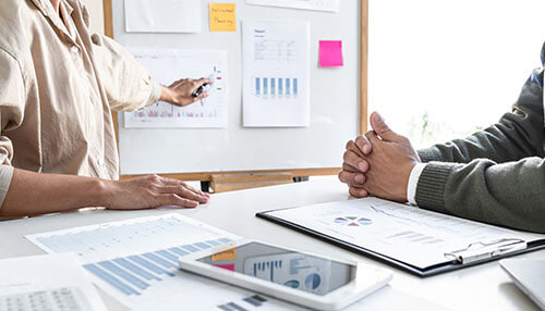 Business capability is the foundation of the strategic planning Two professionals reviewing charts and analytics on a whiteboard and desk, illustrating planning and analysis of business capabilities.