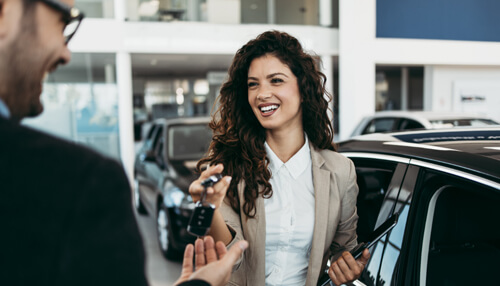 The dealership may be more flexible - tycoonstory | tycoonstory media A smiling woman hands over car keys to a man inside a car dealership, representing in-house auto financing with cars visible in the background.