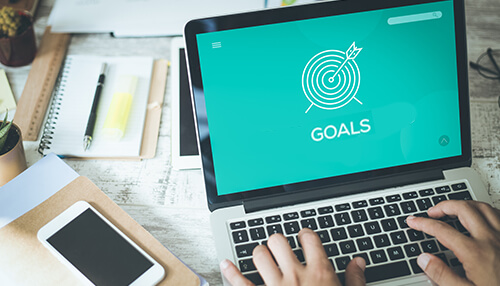 Have clear goals Person typing on laptop displaying goals target graphic on screen at a modern work desk.