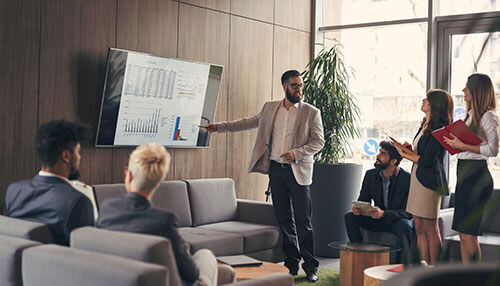 How to create a sales presentation - tycoonstory | tycoonstory media Presenter explaining sales presentation on a screen to colleagues during a team meeting in a modern, collaborative office.
