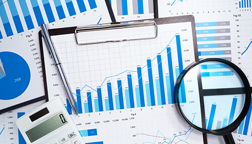 Identify the right level of detail for your data collection efforts Demand planning charts and bar graphs displayed on printed sheets with a magnifying glass, pen, and calculator, showing detailed forecasting analysis.