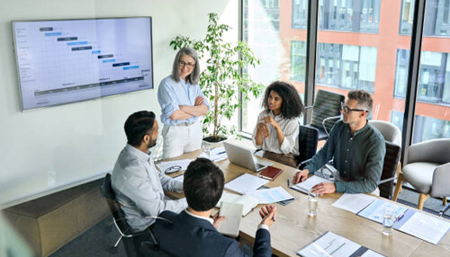 It allows for better employee management Diverse team of business professionals discussing a project schedule on a wall monitor during a meeting, as part of their strategy for effective compensation management.