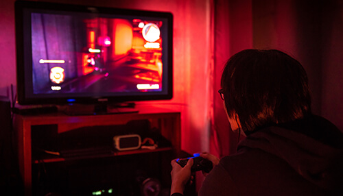 Learning from failure A college student sitting in a dimly lit room playing video games on a large screen, with the glow of the game illuminating the space.