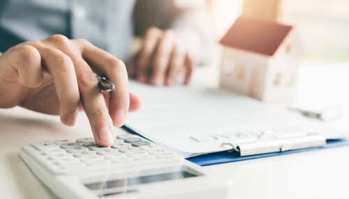 Reasons to buy a home when you have student loans Person using a calculator with paperwork and a small house model, illustrating how to buy a house while dealing with student loans.