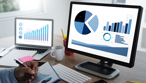 Use business intelligence tools Demand planning data visualizations displayed on computer monitors while a person takes notes, highlighting digital forecasting and analytics.