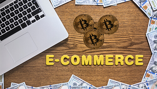 Using crypto for e-commerce - tycoonstory | tycoonstory media Flat lay image showing bitcoin, us dollar bills, and a laptop surrounding the text "e-commerce," symbolizing ecommerce transactions and digital currency.