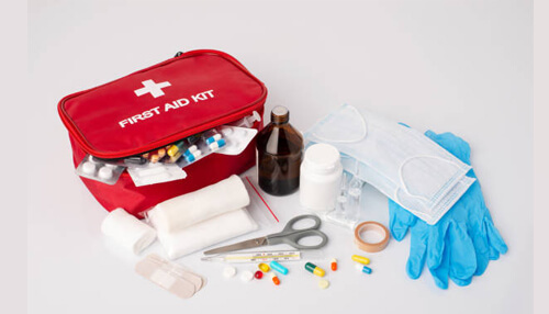 What should be in a first aid kit A top-down view of an open red first aid kit surrounded by medical supplies, including a thermometer, scissors, bandages, pills, face masks, and sterile gloves.