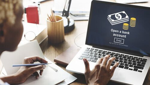 You must have a business bank account Invoice factoring requirement illustrated by a business owner opening a bank account online while managing financial paperwork and cash flow records.