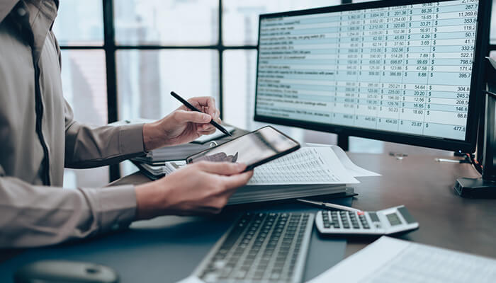 Invoice financing Business professional reviewing financial data on dual monitors while using a tablet and calculator at an office desk.