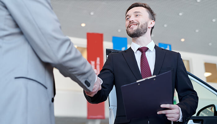 What does a car salesman do - tycoonstory | tycoonstory media Smiling car dealer shaking hands with a customer while holding a clipboard at a car showroom.