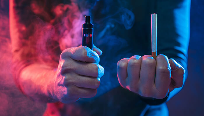Are vapes and cigarettes same - tycoobnstory | tycoonstory media A close-up side view of a hand holding an e-cigarette and a traditional cigarette against a neon red and blue background, comparing modern vaping technology to combustible cigarettes.