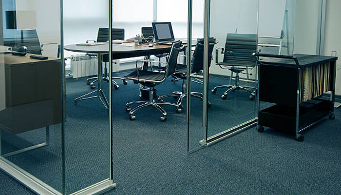 Carpeting A contemporary, glass-walled conference room featuring a round wooden table surrounded by several black, ribbed-leather office chairs on casters. A laptop sits open on the table, and a rolling black utility cart filled with folders is positioned in the foreground to the right. The room has blue carpeting and floor-to-ceiling glass partitions that provide transparency between workspaces.