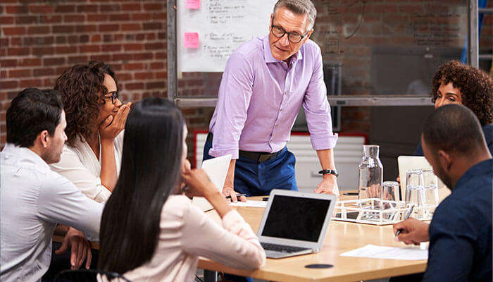 Communicate to your office before you leave A business meeting in progress with a group of professionals discussing ideas around a table, with a leader presenting in front of a whiteboard.