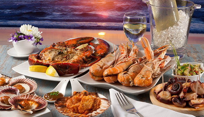 Has wide selection A seafood feast featuring crab, shrimp, scallops, octopus, and other delicacies, set in an elegant dining arrangement with a scenic ocean backdrop.