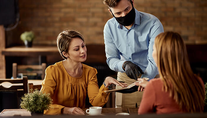 Have the answers - tycoonstory | tycoonstory media Waiter serving customers in a coffee shop as two women review the menu at their table.