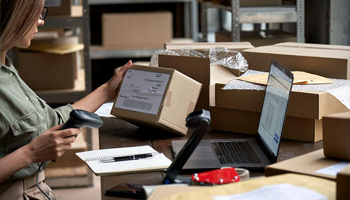 Integrating barcodes into your business A person scanning a barcode on a package in a warehouse setting, with a laptop and notepad on the desk, highlighting the use of barcodes in inventory and shipping management.