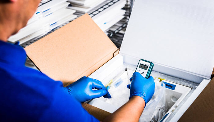 The main types of temperature-controlled packaging Person handling a shipment using temperature-controlled packaging, checking the temperature with a digital device while wearing protective gloves.