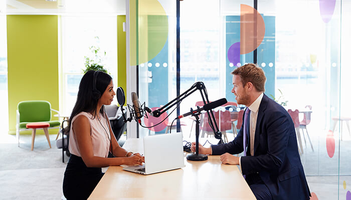 Eight steps to create podcasts for your business in 2023 - tycoonstory | tycoonstory media A man and a woman in professional attire recording a podcast or interview, sitting at a table with microphones, a laptop, and headphones in a brightly lit modern office setting.