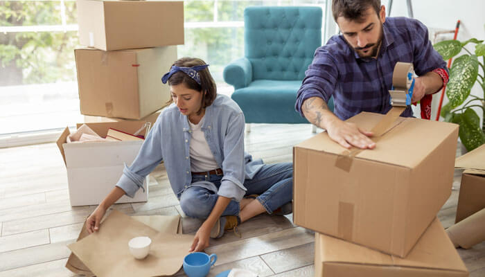 Not packing properly A young couple is packing for relocating, with the woman wrapping fragile cups and the man sealing a large cardboard box with tape.