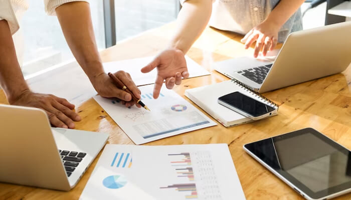 Primary market research Team collaborating on desk-based market research, reviewing charts and data sheets on a wooden desk with laptops, tablet, and smartphone.