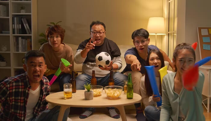 Social interaction Group of friends excitedly watching a sports game at home, cheering with colorful noisemakers and a soccer ball, enjoying snacks and drinks.