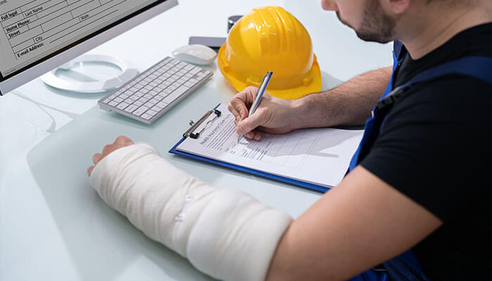 Workers compensation insurance A worker with a bandaged arm filling out an injury report form after an accident at the workplace, with a hard hat beside him.