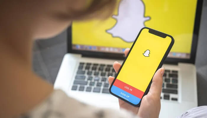 Snapchat-tycoonstory | tycoonstory media Person holding a smartphone with the snapchat login screen open, with a laptop showing the snapchat logo in the background, highlighting snap my ai use.