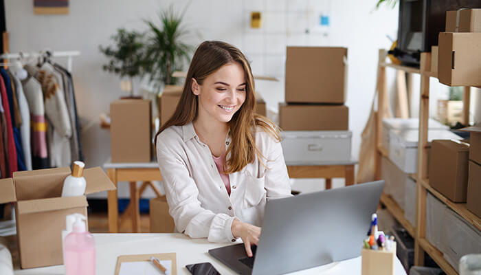 Dropshipping-tycoonstory | tycoonstory media A young woman working on her laptop in a home office, managing her dropshipping business in the e-commerce industry, with packing boxes visible in the background.