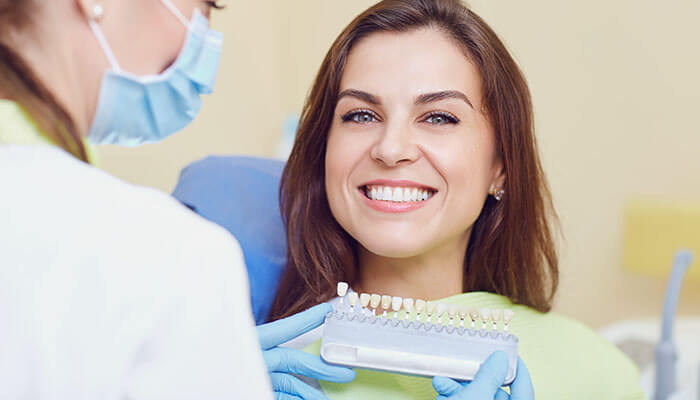 Disadvantages of dental crowns A smiling female patient sitting in a dentist's chair, looking at the camera while a dentist (wearing a mask and gloves) holds up a tooth shade guide. This illustrates the color selection for treatments such as Dental Crowns or veneers.