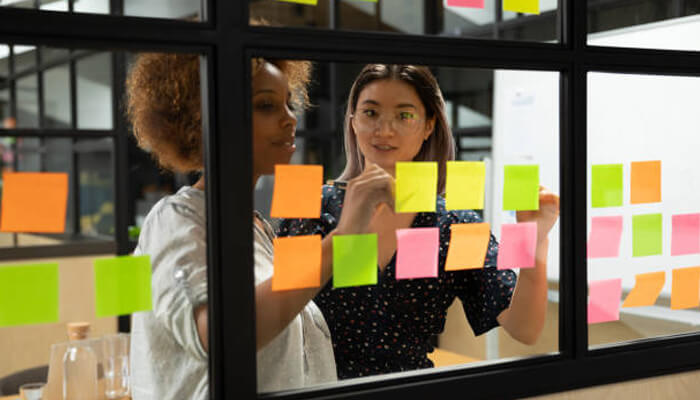Identify purpose Two women collaborating in an office environment, brainstorming and arranging colorful sticky notes on a window, representing the planning phase for a tiny business idea