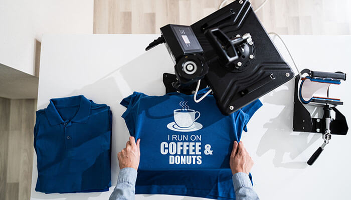 Print-on-demand t-shirt production process with heat press machine printing custom coffee and donuts design on blue apparel.