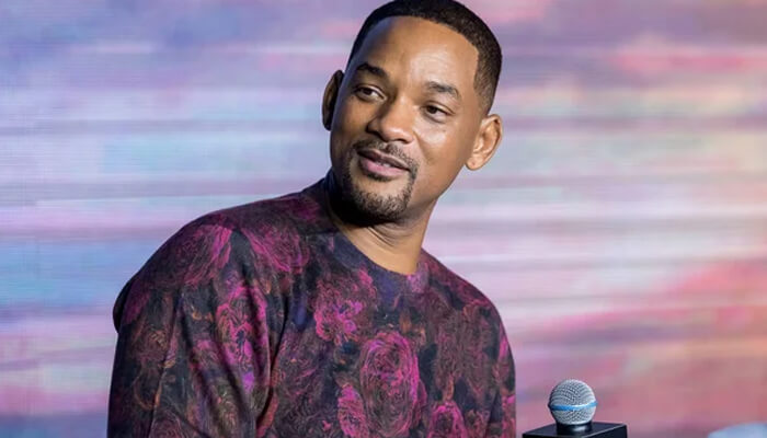 Will smith's Will smith’s smiling portrait as he speaks into a microphone, wearing a patterned maroon sweater against a colorful blurred background.