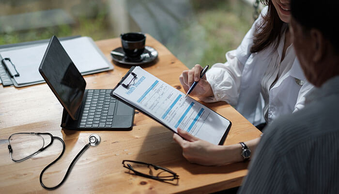 Additional benefits and riders Life insurance consultation scene where a professional reviews and fills out insurance forms with a client at a desk, alongside a laptop and stethoscope.