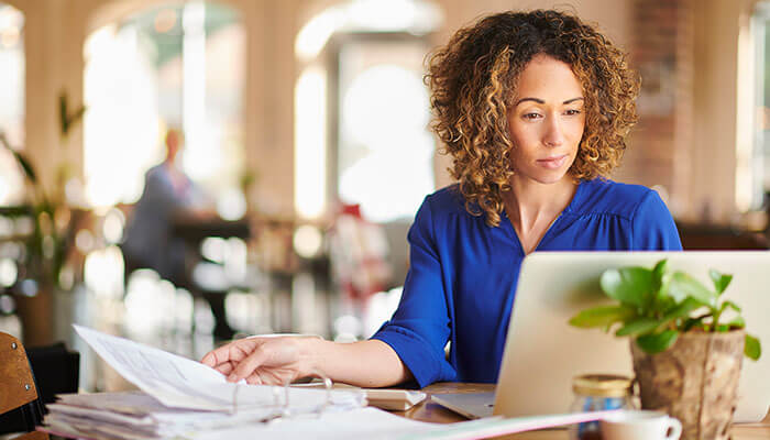 Wfh A woman working on a laptop in a café, reviewing documents while sitting at a table surrounded by papers and a small potted plant.