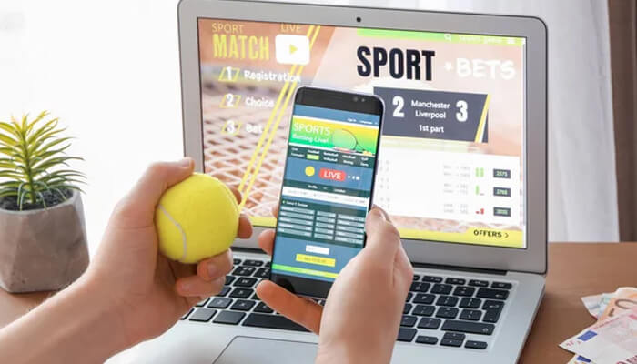 Here are five starting pointers to identifying sports betting value - tycoonstory | tycoonstory media A person holding a smartphone and tennis ball while using a laptop to place sports betting bets on live sports matches through an online platform.