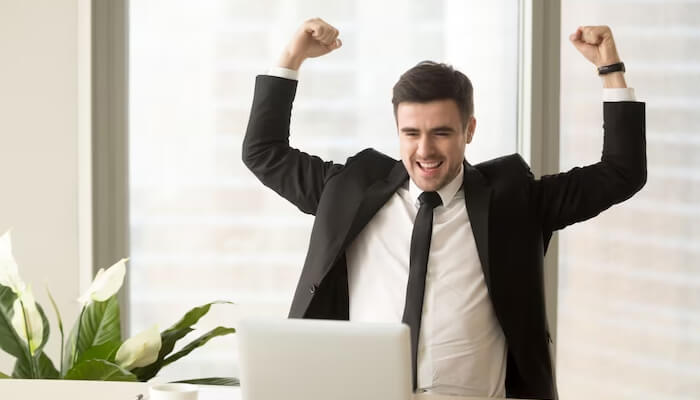 Self-motivated Successful businessman celebrating at a laptop in an office, raising his fists in victory, representing the achievement and growth journey of a secret entrepreneur.