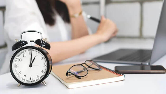 Job skills A workspace with a classic alarm clock, eyeglasses, notebook, and laptop, showing a person working in the background.