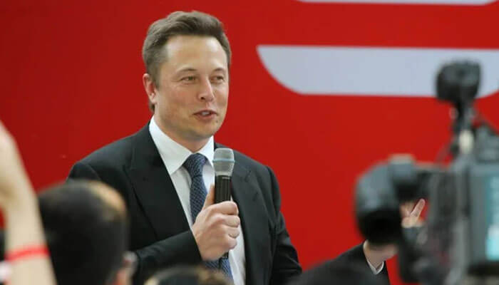 Mr musk - tycoonstory | tycoonstory media Elon musk speaking at an event with a microphone, dressed in a suit, addressing an audience with cameras capturing the moment.