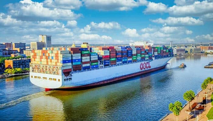 The-shifts-tycoonstory | tycoonstory media A large cargo ship loaded with colorful containers, sailing along a waterway with city buildings in the background and a bright, cloudy sky.