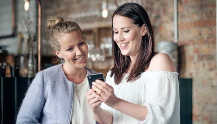 Use comparison websites - tycoonstory | tycoonstory media Two women smiling while looking at a smartphone together in a cozy indoor setting.