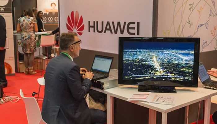 Huawei Man in a suit working on a laptop at a huawei exhibit booth, with a large screen showing a city at night.