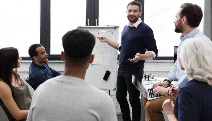 Improved communication A business professional presenting business systems and workflow strategies on a whiteboard to a diverse team during a meeting in a modern office.